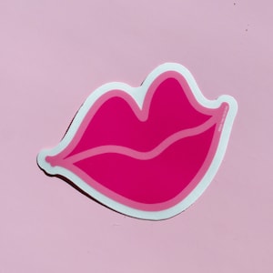 Puede incluir: Una pegatina rosa y blanca con forma de labio de dibujos animados.