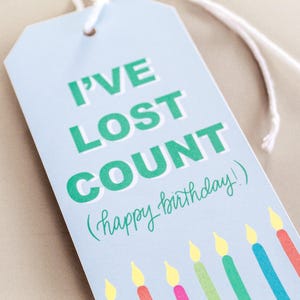 May include: A pale blue gift tag with the words "I've lost count (happy birthday!)" in green, and a row of colourful candles. The tag is rectangular with a hole at the top and a white string.