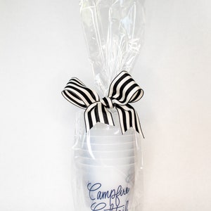May include: A set of clear plastic cups with a black and white striped ribbon tied around them. The cups are wrapped in clear plastic and have the text "Campfire Cocktail" printed on them in blue.