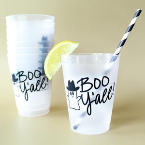 May include: Clear plastic cups with a Halloween-themed design. The cups feature a ghost wearing a hat and the phrase "BOO Y'all!" in black script. One cup has a lemon slice and a black and white striped straw.