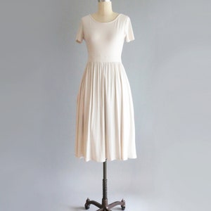 May include: A simple, off-white, short-sleeved dress with a gathered skirt. The dress is on a mannequin and is displayed against a light gray background.