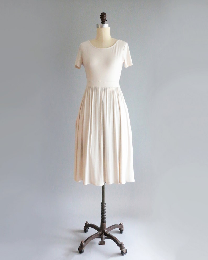 May include: A simple, off-white, short-sleeved dress with a gathered skirt. The dress is on a mannequin and is displayed against a light gray background.