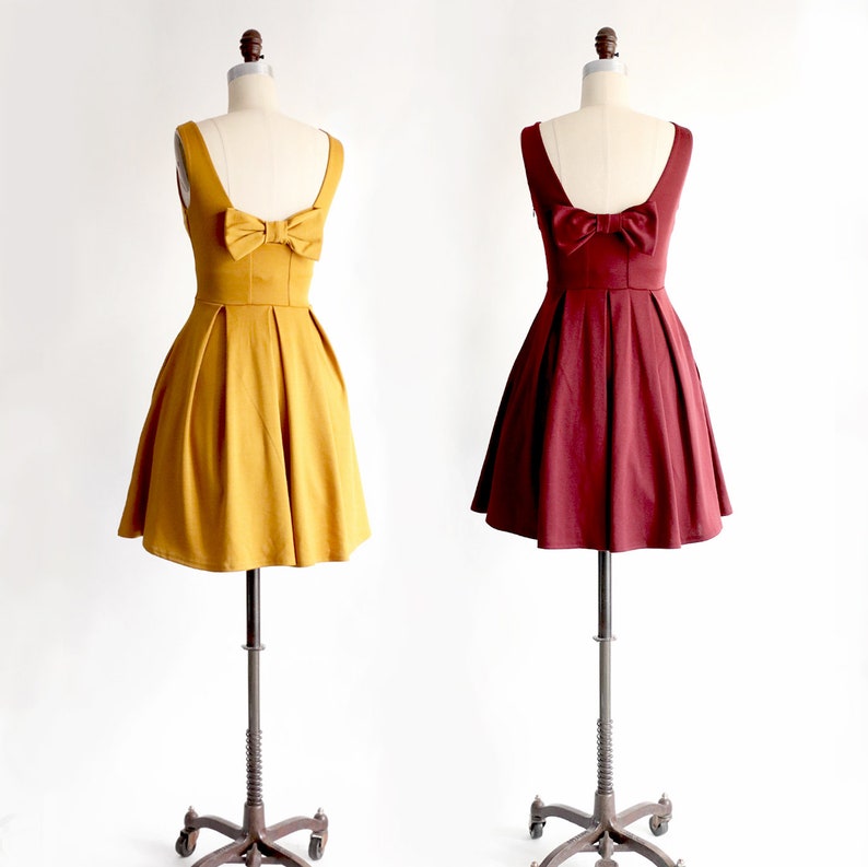 May include: Two sleeveless dresses on mannequins. One is mustard yellow, the other is burgundy. Both have a bow detail on the back and a pleated skirt. The dresses are displayed on adjustable metal stands with wheels.