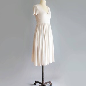 May include: A simple, off-white, knee-length dress with a V-neckline and short sleeves. The dress is made of a soft, flowing fabric and has a gathered skirt.