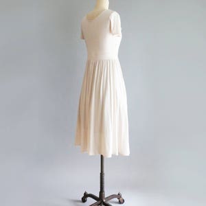 May include: A simple, off-white, short-sleeved dress with a gathered skirt. The dress is on a mannequin with a metal stand.