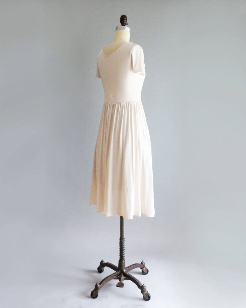 May include: A simple, off-white, short-sleeved dress with a gathered skirt. The dress is on a mannequin with a metal stand.