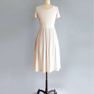 May include: A simple, off-white, knee-length dress with short sleeves and a gathered skirt. The dress is on a dress form with a brown metal stand.