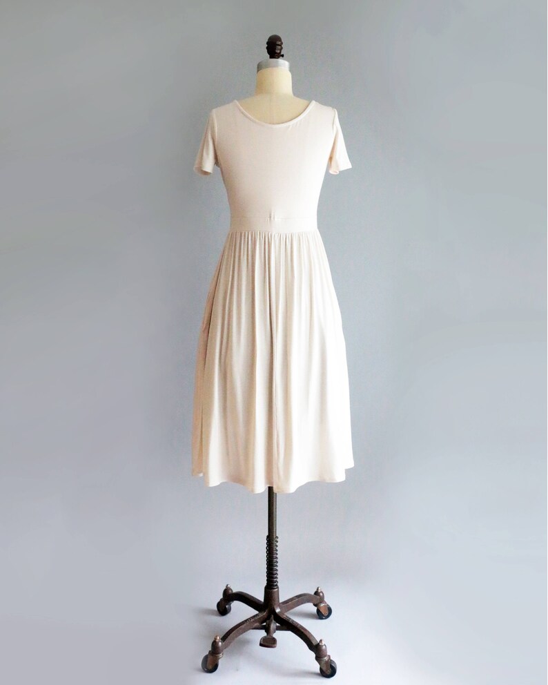 May include: A simple, off-white, knee-length dress with short sleeves and a gathered skirt. The dress is on a dress form with a brown metal stand.