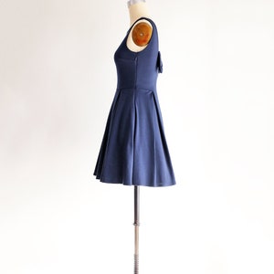 May include: A navy blue sleeveless dress with a pleated skirt and a small bow detail on the back. The dress is displayed on a mannequin, showcasing its fit and design. The dress is made of a smooth, solid fabric.