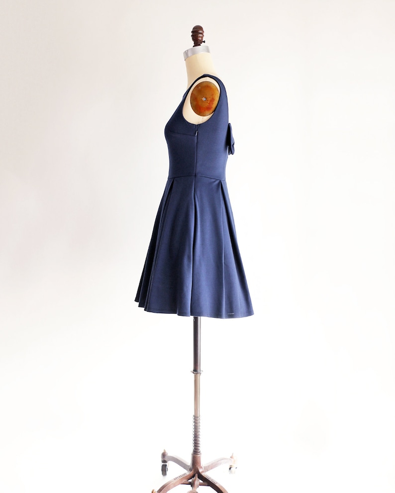 May include: A navy blue sleeveless dress with a pleated skirt and a small bow detail on the back. The dress is displayed on a mannequin, showcasing its fit and design. The dress is made of a smooth, solid fabric.