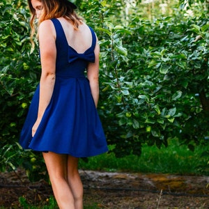 May include: A woman in a royal blue dress with a bow detail on the back, and a black hat, stands in an orchard. The dress has a flared skirt. The background features lush green foliage and a dirt path.