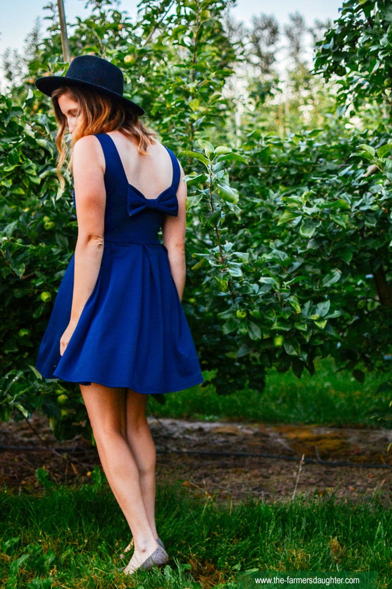May include: A woman in a royal blue dress with a bow detail on the back, and a black hat, stands in an orchard. The dress has a flared skirt. The background features lush green foliage and a dirt path.