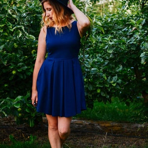 May include: A sleeveless, navy blue dress with a pleated skirt. The dress is paired with a black hat. The image is set in an orchard with green foliage and grass.
