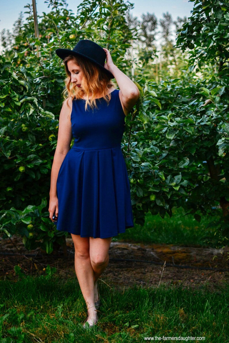 May include: A sleeveless, navy blue dress with a pleated skirt. The dress is paired with a black hat. The image is set in an orchard with green foliage and grass.
