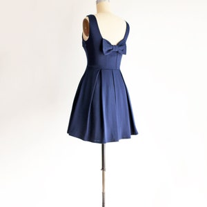 May include: A navy blue dress displayed on a mannequin. The dress features a scoop neckline, a fitted bodice, and a flared skirt. A bow detail adorns the back. The dress is sleeveless and appears to be made of a smooth, flowing fabric.