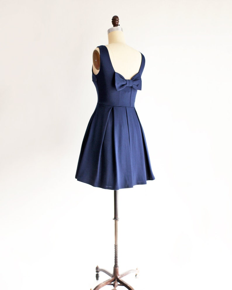 May include: A navy blue dress displayed on a mannequin. The dress features a scoop neckline, a fitted bodice, and a flared skirt. A bow detail adorns the back. The dress is sleeveless and appears to be made of a smooth, flowing fabric.