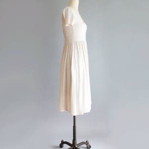 May include: A simple, off-white, knee-length dress with a V-neckline and short sleeves. The dress is on a dress form with a metal stand.