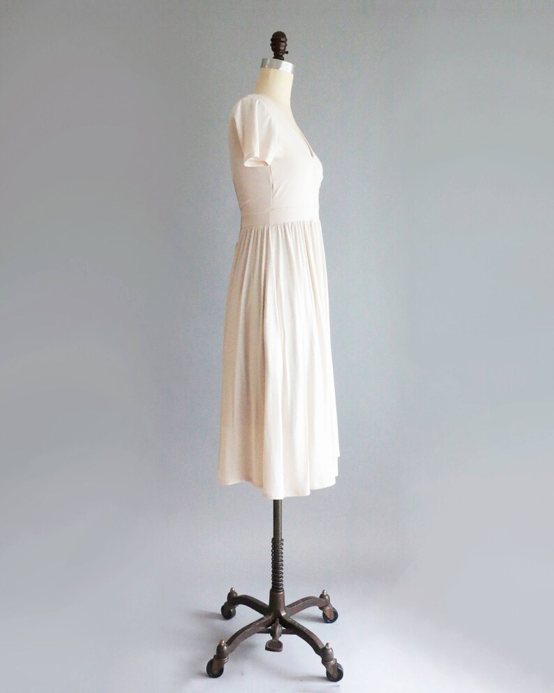 May include: A simple, off-white, knee-length dress with a V-neckline and short sleeves. The dress is on a dress form with a metal stand.