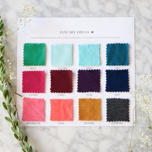May include: A color swatch card for the "January Dress" featuring samples in various shades, including Kelly green, fuchsia, and mustard. The card is displayed on a marble surface, with sprigs of greenery and white flowers.
