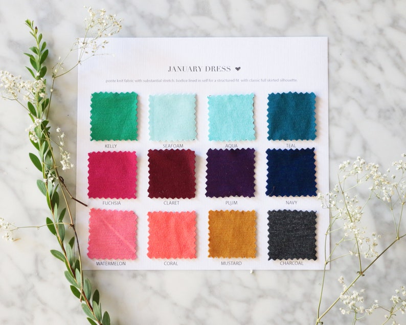 May include: A color swatch card for the "January Dress" featuring samples in various shades, including Kelly green, fuchsia, and mustard. The card is displayed on a marble surface, with sprigs of greenery and white flowers.