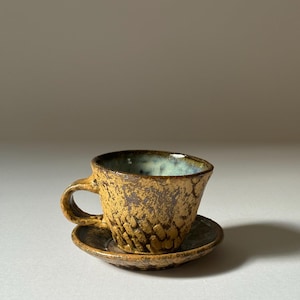 May include: A small, yellow ceramic teacup and saucer set with a textured, speckled glaze.