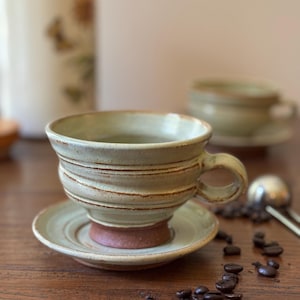 Puede incluir: Una taza de té y un platillo de cerámica verde claro con un borde marrón. La taza tiene una forma redondeada y un asa. El platillo es ligeramente más grande que la taza y tiene un diseño a juego. Granos de café esparcidos sobre una superficie de madera.