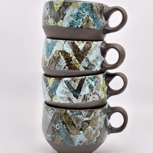 May include: Four ceramic mugs stacked. Each mug has a dark brown base and handle. The mugs feature a textured pattern in blue, green, and brown. The mugs are stacked against a white background.