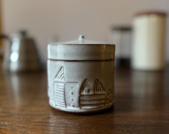 Puede incluir: Un tarro de cerámica blanco con tapa, que presenta un diseño pintado a mano de un pequeño pueblo con casas y árboles. El tarro tiene una superficie ligeramente texturizada.