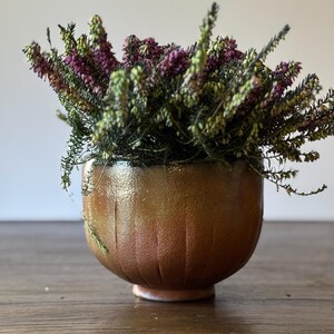 May include: A small, round, terracotta-colored ceramic pot with a textured surface. The pot is filled with a bouquet of pink and green heather.