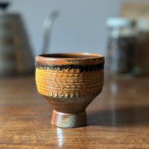 May include: A brown ceramic cup with a textured surface and a black band around the top. The cup is sitting on a wooden table.