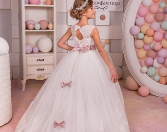 May include: A white formal dress with a tulle skirt and lace trim. The dress features a pink sash and bows, and a keyhole back. The girl is wearing a jeweled headband. The background includes a white cabinet and wall decor.