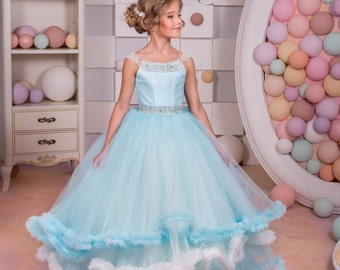 Può includere: Una bambina che indossa un abito di tulle azzurro chiaro con volant bianchi. L'abito ha una cintura con perline e una scollatura con perline. La bambina è in piedi davanti a un muro bianco con una cornice bianca attorno a un muro di palline colorate.