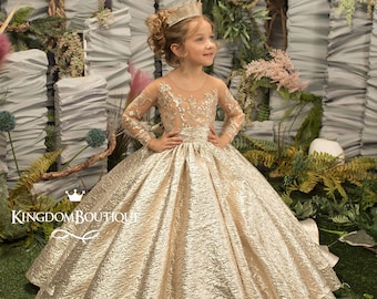 May include: A young girl wearing a gold, sparkly, tulle dress with long sleeves and a crown. The dress has a fitted bodice and a full skirt. The girl is standing in front of a backdrop of white and green foliage.