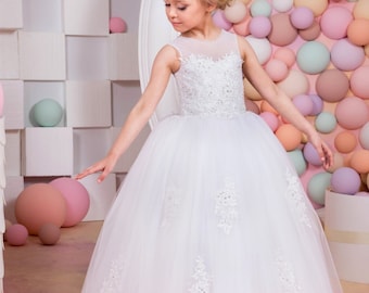 Pode incluir: Um vestido branco de tule com detalhes em renda e um corpete transparente. O vestido é usado por uma menina com cabelo loiro e uma tiara. O fundo é uma parede de balões de cores pastel.