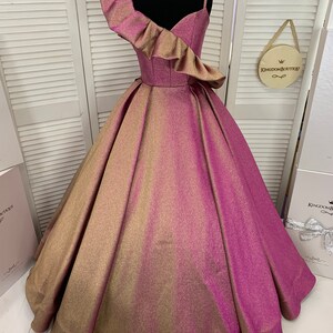 May include: A gold and pink ombre ball gown with a single ruffle shoulder strap. The dress has a fitted bodice and a full skirt.