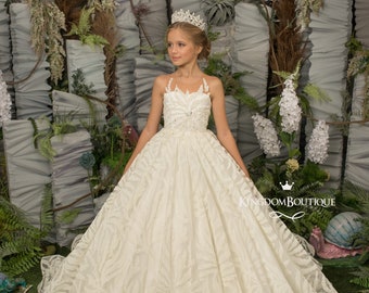 May include: A young girl wearing a white tulle dress with a floral pattern and a sparkly bodice. She is wearing a tiara and standing in front of a green and gray background.