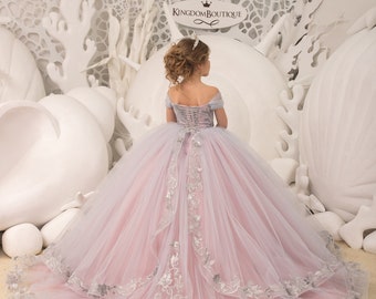 May include: A young girl wearing a pink and gray tulle ball gown with silver embroidery. The gown has a sweetheart neckline and off-the-shoulder straps. The girl is standing in front of a white background with white decorative elements. The text "Kingdom Boutique" is visible in the image.