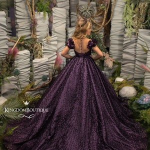 May include: A purple ball gown with a sheer bodice and floral embellishments. The gown has a full skirt and is made of a sparkly fabric. The model is standing in front of a backdrop of greenery and white fabric.