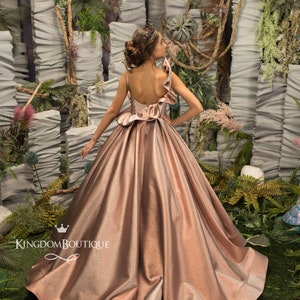 May include: A rose gold sparkly princess dress with a ruffled bodice and a full skirt. The dress is photographed against a backdrop of white and green foliage. The text "KingdomBoutique" is visible on the dress.