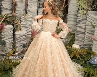 May include: A young girl wearing a long, white, lace dress with a sheer bodice and long sleeves. The dress has a floral pattern and a belt around the waist. The girl is standing in front of a backdrop of green plants and white fabric.