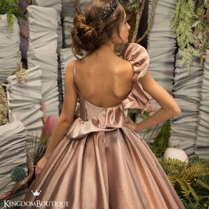 May include: A rose gold sleeveless dress with a ruffled bodice and a full skirt. The dress has a low back and is accented with a sparkly fabric. The dress is photographed against a backdrop of white and green foliage.