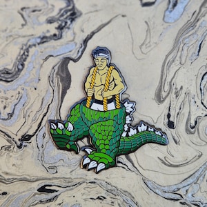 May include: A colorful enamel pin featuring a muscular figure with a headband and rope, emerging from a green Godzilla-like creature. The pin has a black outline and is set against a marbled background.