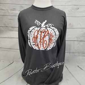 May include: A charcoal long-sleeved top with a white pumpkin design and an orange monogram. The pumpkin has a distressed appearance. The text "Rooster Boutique" is written in cursive at the bottom of the shirt.