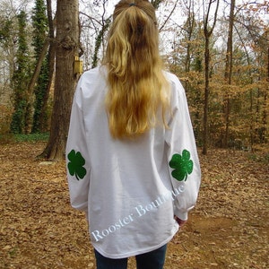 May include: White long-sleeved shirt with two green glitter shamrocks on the sleeves. The text "Rooster Boutique" is printed in white on the lower back of the shirt. The shirt is worn with blue jeans.