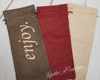 May include: Three burlap wine bottle bags in brown, red, and beige. The brown bag has the word "enjoy" printed in a light color. Each bag has a drawstring closure at the top. The bags are made of a textured fabric.