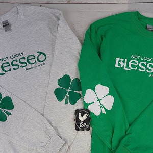 May include: Two long-sleeved shirts, one gray and one green, with the words "NOT LUCKY BLESSED Romans 4:7-8" printed on the front. Each sleeve features a four-leaf clover design. The shirts are displayed on a light wood surface.