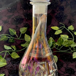 May include: A clear glass decanter with a decorative stopper. The decanter has a pear-shaped body and a diamond-cut base. The stopper is a square shape with a diamond pattern. The glass has an iridescent finish, reflecting colors of purple, gold, and green.
