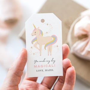 Puede incluir: Una etiqueta de regalo blanca con una ilustración de unicornio rosa y purpurina dorada. La etiqueta dice "You made my day magical! Love, Hazel."