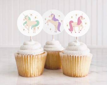 May include: Three cupcakes with white frosting, each topped with a round unicorn-themed decoration. The unicorn illustrations feature pastel colors, including pink, purple, and mint green, with gold star accents. The cupcakes sit on a white surface.
