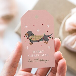 May include: Pink Christmas gift tag with a cartoon dachshund wearing a plaid scarf and holding Christmas lights. The text reads "Merry Christmas Love, The Pacy's".